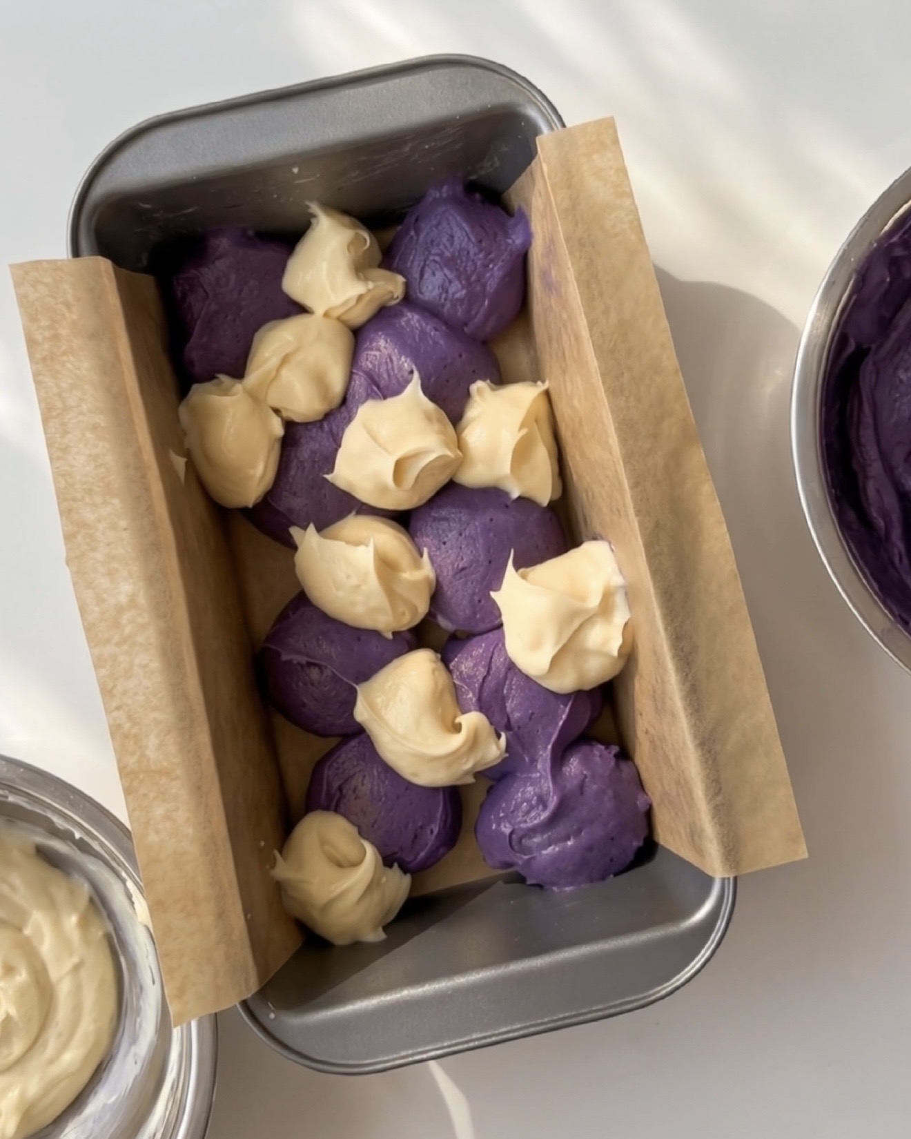 Cuillerées de pâte d'ube violette et de pâte à la vanille alternées dans un moule à cake chemisé pour le cake marbré ube Ube Nolia