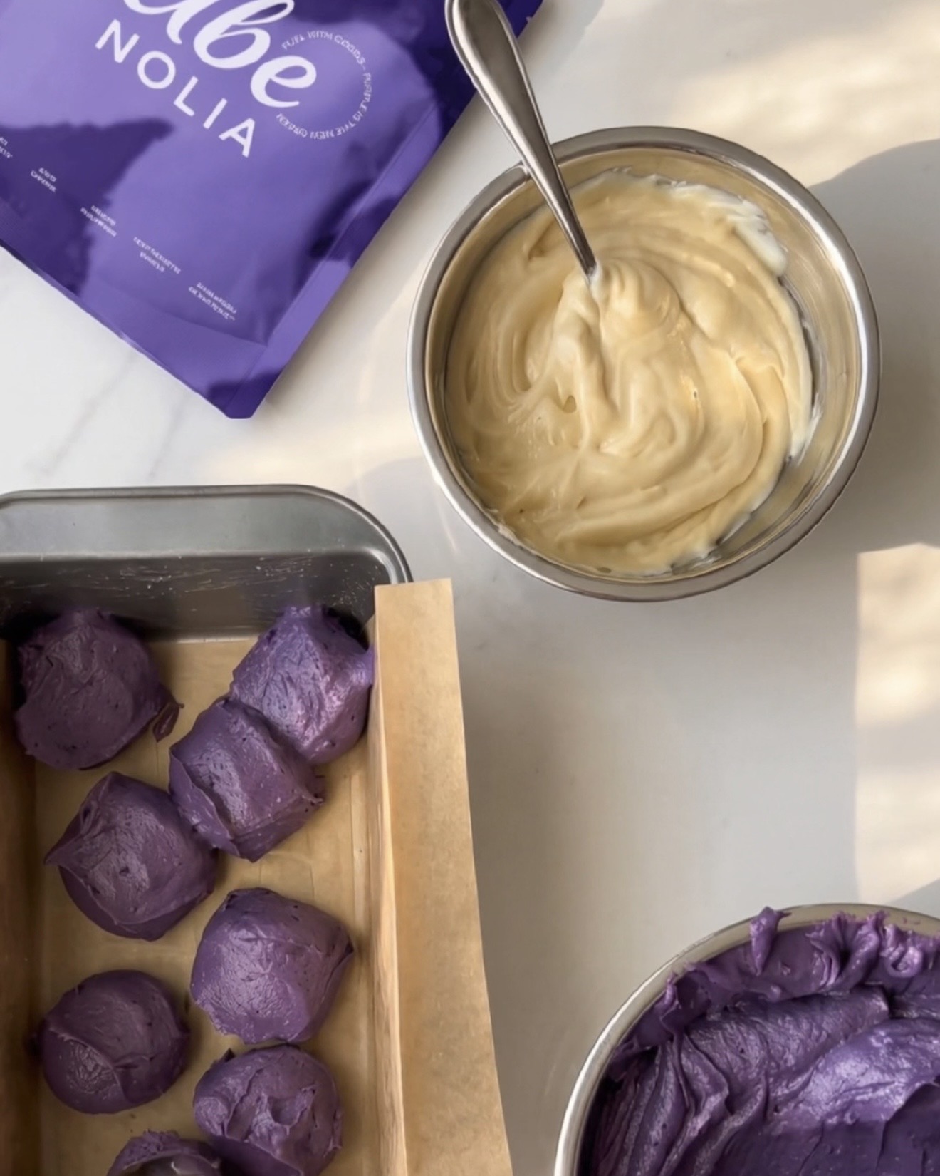 Ube halaya fait maison, pâte à la vanille et cuillerées de pâte d'ube violette dans un moule à cake, à côté d'un sachet de poudre d'ube Ube Nolia