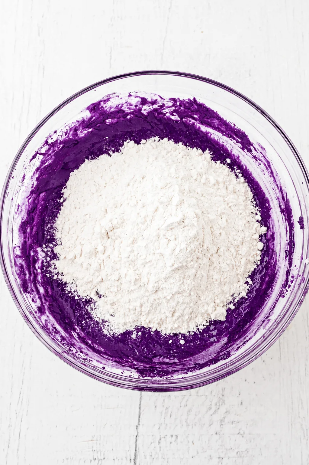 Farine ajoutée sur la pâte à brownie ube au chocolat blanc violet foncé dans un bol en verre, prête à être incorporée avec de l'UBE NOLIA® powder