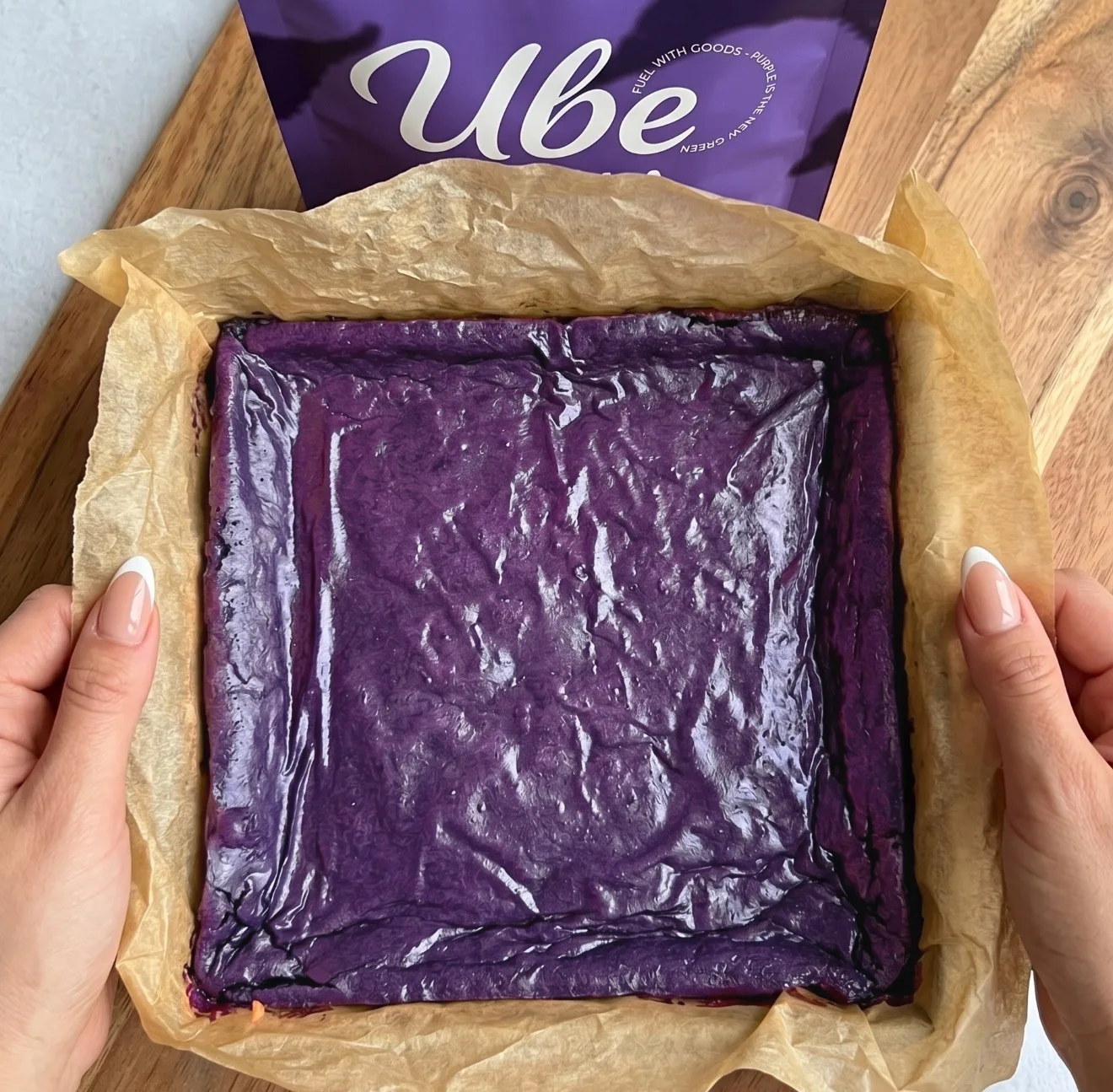 Pâte à brownie ube violet foncé superposée dans un moule carré tapissé de papier sulfurisé à côté d'un sachet d'UBE NOLIA® powder